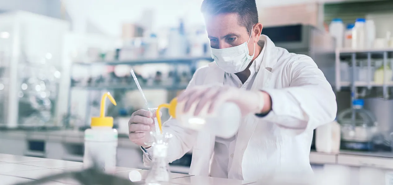 A scientist is carefully pouring liquid from a bottle into a container in a laboratory setting.
